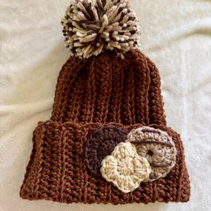 May include: A brown, handmade crochet beanie with a large pom-pom on top. The beanie features three decorative crochet appliques in shades of brown and beige. The word "Walnut" is printed at the bottom.