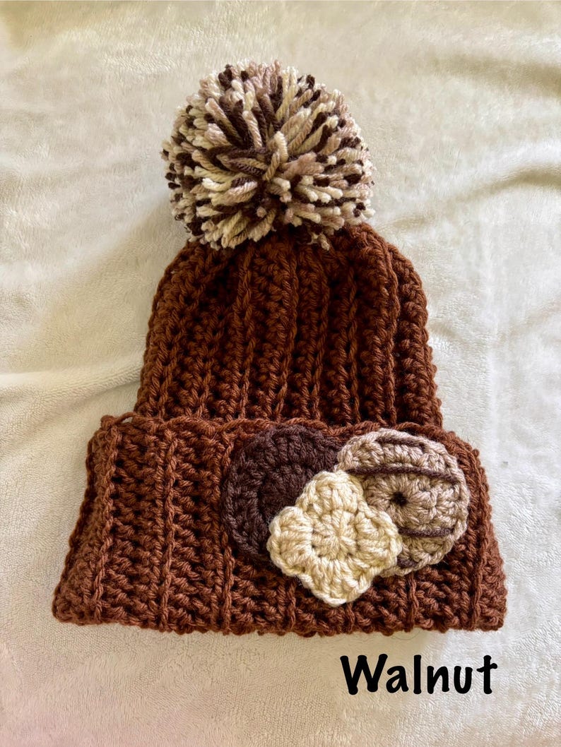 May include: A brown, handmade crochet beanie with a large pom-pom on top. The beanie features three decorative crochet appliques in shades of brown and beige. The word "Walnut" is printed at the bottom.