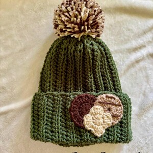May include: A green, ribbed-knit beanie with a large, multi-toned pom-pom on top. The hat features three crocheted floral embellishments in shades of brown and beige. The text "Deep Forrest" is printed at the bottom.