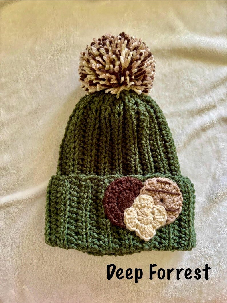 May include: A green, ribbed-knit beanie with a large, multi-toned pom-pom on top. The hat features three crocheted floral embellishments in shades of brown and beige. The text "Deep Forrest" is printed at the bottom.