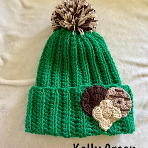 May include: A Kelly Green crochet beanie with a brown and tan pom-pom on top. The hat features a brown and tan cookie applique on the front. The text "Kelly Green" is displayed on the bottom.