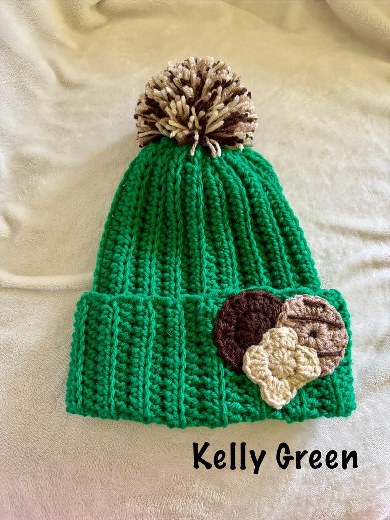 May include: A Kelly Green crochet beanie with a brown and tan pom-pom on top. The hat features a brown and tan cookie applique on the front. The text "Kelly Green" is displayed on the bottom.