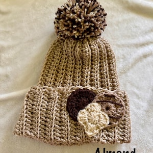 May include: A handmade, crochet beanie in almond color with a large pom-pom on top. The brim features three decorative crochet appliques in shades of brown and beige. The word "Almond" is printed on the bottom of the image.