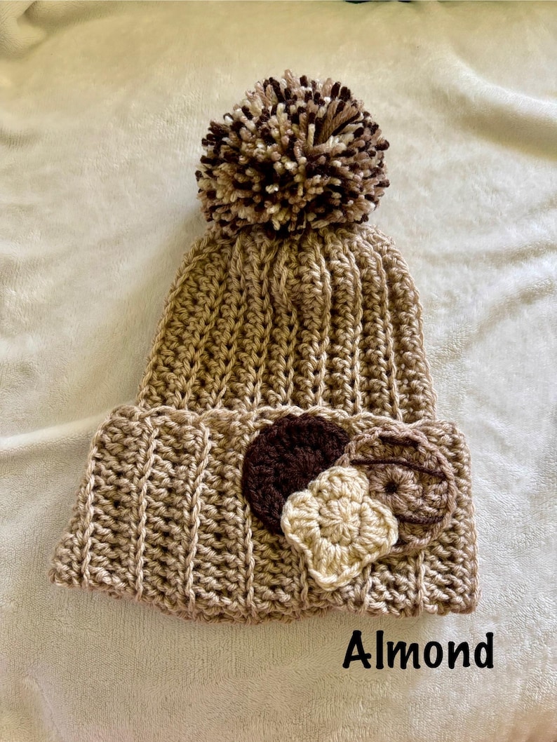 May include: A handmade, crochet beanie in almond color with a large pom-pom on top. The brim features three decorative crochet appliques in shades of brown and beige. The word "Almond" is printed on the bottom of the image.