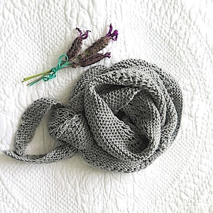 May include: A gray knitted scarf with a loose, open weave. The scarf is folded and arranged on a white textured surface. A small bouquet of purple lavender flowers is placed to the side of the scarf.