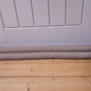 May include: A beige fabric door draft excluder, designed to block draughts and insulate. The cylindrical shape is visible against a white door and wooden floor. The excluder is approximately 91 cm long.