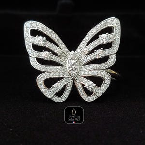 May include: A silver butterfly ring with a detailed design. The ring is encrusted with small, clear gemstones, creating a sparkling effect. The butterfly's wings are open, and the ring is set against a black background.