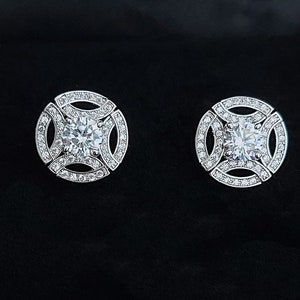 Può includere: Due orecchini a lobo argentati con un design rotondo e geometrico. Ogni orecchino presenta una grande pietra centrale rotonda e trasparente, circondata da pietre più piccole e scintillanti. Gli orecchini sono su sfondo nero.