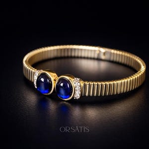 Peut inclure: Bracelet doré avec un design côtelé. Le bracelet présente deux grandes pierres précieuses rondes bleu foncé serties dans des lunettes dorées, accentuées de petites pierres claires. Le bracelet est présenté sur un fond noir.