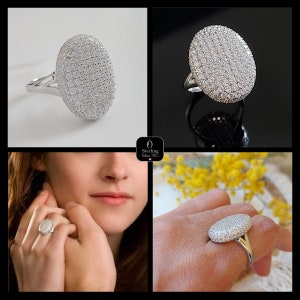 Puede incluir: Un anillo de plata con una piedra central de forma ovalada. La piedra está cubierta de pequeñas piedras de circonita cúbica transparentes. El anillo se muestra en el dedo de una modelo y sobre un fondo blanco.