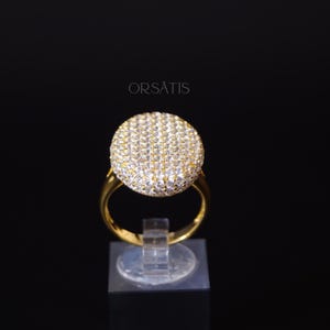 May include: A gold-colored ring with a large, round, sparkling centerpiece. The centerpiece is covered in small, clear stones. The ring is displayed on a clear stand against a black background. The word "ORSATIS" is printed in white.