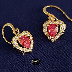 May include: A pair of gold-colored heart-shaped earrings. Each earring features a teardrop-shaped red gemstone surrounded by small, clear crystals. The earrings have a hook closure and are set against a dark blue background.