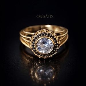 Peut inclure: Bague en or avec une grosse pierre précieuse ronde et transparente au centre, entourée de petites pierres noires. La bague a un anneau détaillé et le mot "ORSATIS" est au-dessus.