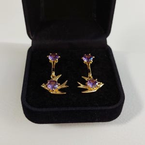Peut inclure: Une paire de boucles d'oreilles dorées avec un motif d'oiseau et des pierres précieuses violettes. Les boucles d'oreilles sont présentées dans un écrin noir avec un intérieur en velours. Elles sont de type pendantes.