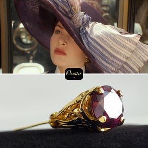 Peut inclure: Une femme porte un grand chapeau violet avec un nœud rayé. En dessous, une épingle dorée au design orné tient une grosse pierre précieuse rouge à facettes. L'épingle est posée sur un fond noir.