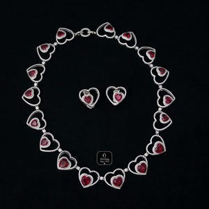 May include: A heart-shaped necklace and matching earrings set. The jewelry features a series of open heart designs, each with a vibrant red center stone and outlined with small, sparkling accents. The set is displayed on a black background.