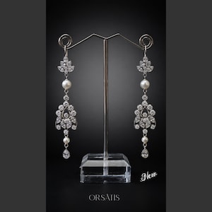 Peut inclure: Une paire de boucles d'oreilles chandelier argentées avec des cristaux facettés transparents et des perles blanches. Le design comprend une section supérieure florale, un motif central orné et une goutte de cristal. Texte ORSÂTIS et New.
