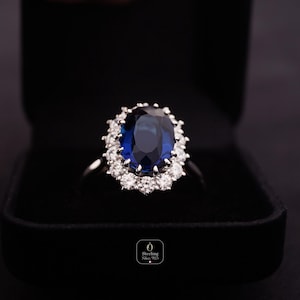 Könnte beinhalten: Ein silberner Ring mit einem großen, ovalen, tiefblauen Edelstein, umgeben von kleineren weißen Steinen. Der Ring wird in einer schwarzen Samt-Ringbox präsentiert. Ein klassisches Design für besondere Anlässe.