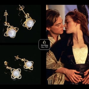 Peut inclure: Boucles d'oreilles pendantes dorées avec un centre en forme de perle et un motif floral. Les boucles d'oreilles sont présentées sur un fond noir. L'image comprend également une scène du film Titanic.