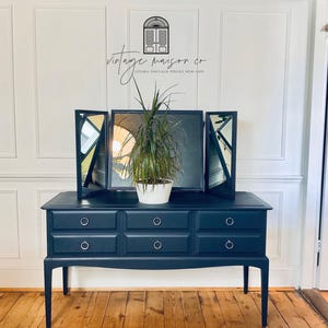 May include: A dark navy blue vanity table featuring six drawers with silver ring pulls and a matching tri-fold mirror. A green plant in a white pot sits on the centre of the surface. The logo for vintage maison co is visible above.