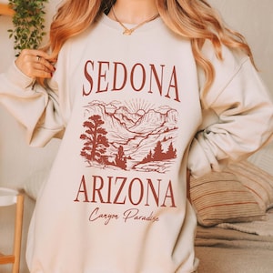 May include: A beige sweatshirt with a red graphic print of a mountain landscape and the text "Sedona Arizona Canyon Paradise".