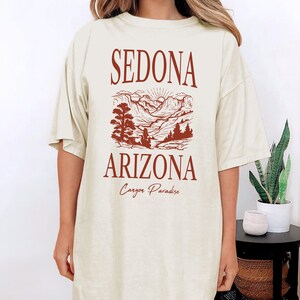 May include: A beige oversized t-shirt with a graphic print of a mountain landscape in Sedona, Arizona. The text "Sedona Arizona Canyon Paradise" is printed in brown on the shirt.