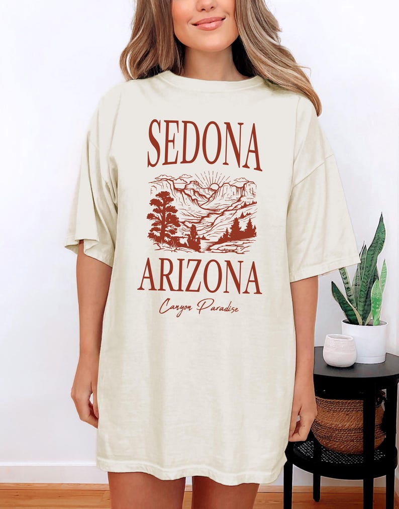 May include: A beige oversized t-shirt with a graphic print of a mountain landscape in Sedona, Arizona. The text "Sedona Arizona Canyon Paradise" is printed in brown on the shirt.