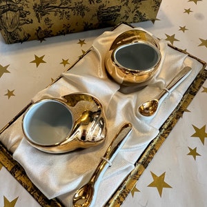May include: Two gold-rimmed porcelain bowls with spoons in a white satin box with a gold patterned border. The box is on a white background with gold stars.