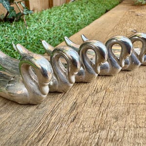 May include: A row of silver swan-shaped napkin rings. The swan's necks curve gracefully, and the bodies are detailed. The napkin rings are arranged on a weathered wooden surface, with green grass and foliage in the background.