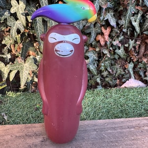 Könnte beinhalten: Ein braunes, affenförmiges Spielzeug mit einem lächelnden Gesicht und einem regenbogenfarbenen Vogel oben drauf. Das Spielzeug steht auf einer Holzoberfläche, mit einem Hintergrund aus grünem Efeu.