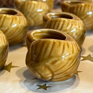 May include: A group of small, tan ceramic containers with a textured, swirling pattern. Each has a rounded body and a small handle. The containers are arranged on a surface with gold star-shaped decorations.