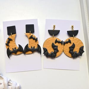 May include: Two pairs of earrings on white display cards. The first pair features black square studs with yellow crescent moons and black bats. The second pair features black semi-circle studs with yellow circular discs and black bats.