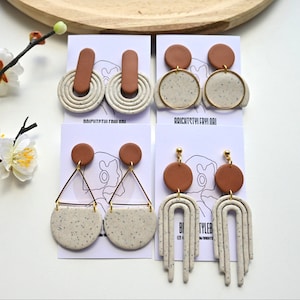 May include: Three pairs of handmade earrings with brown and white speckled clay. The earrings are in different styles, including a circle, a triangle, and a curved shape.