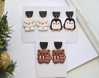 May include: A collection of handmade polymer clay earrings featuring winter-themed designs. The earrings include snowmen, penguins, and brown bears wearing red and white striped scarves. Each pair has a black top and is displayed on a white card.
