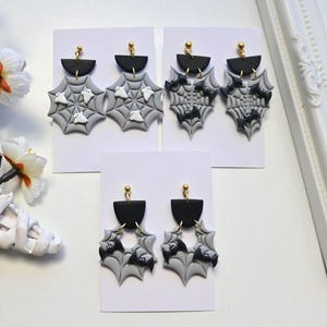 Pode incluir: Três pares de brincos pendentes com topos em semicírculo preto e pingentes cinzentos em forma de teia de aranha. Um par exibe figuras de fantasmas brancos, enquanto os outros dois pares apresentam figuras de morcegos pretos com pinos dourados.