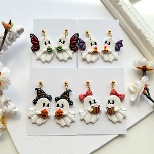 Può includere: Una collezione di orecchini fatti a mano a tema Halloween. Gli orecchini presentano figure di fantasmi bianchi con vari accessori, tra cui cappelli da strega, zucche, libri, fiocchi e ali. Ogni paio ha un gancio color oro.