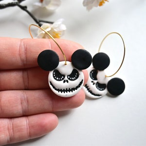 May include: A pair of hoop earrings featuring a white circular charm with a black stitched mouth and eye details, topped with two black circular ears. The charms are attached to thin gold-toned metal hoops, creating a character-inspired design.