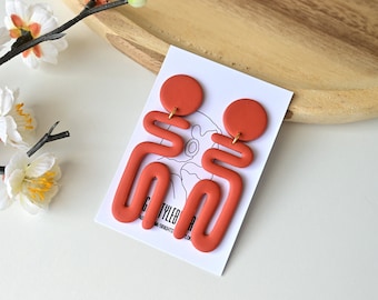 May include: A pair of orange abstract dangle earrings with a geometric design. The earrings are made of polymer clay and have a matte finish.