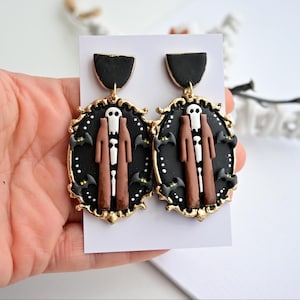 Pode incluir: Um par de brincos pendentes com uma moldura oval preta e bordas douradas. Dentro de cada moldura, uma figura veste um casaco castanho comprido e uma máscara de caveira branca, rodeada por pequenos morcegos pretos e pontos brancos num fundo preto.