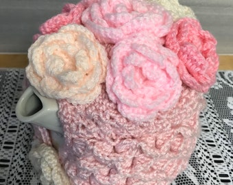 Può includere: Un copri-teiera in crochet rosa con un design di rose in crochet rosa, pesca e bianche.