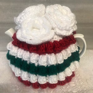 May include: White crocheted teapot cozy with red and green stripes. The cozy has three white crocheted flowers on top.