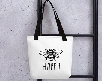 May include: White tote bag with black handles, featuring a black and white illustration of a bee and the word "HAPPY" in a handwritten font. The bag is hanging against a gray speckled background.