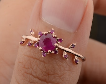 May include: A rose gold ring with a ruby gemstone surrounded by smaller ruby stones. The ring features a delicate branch design with leaves.