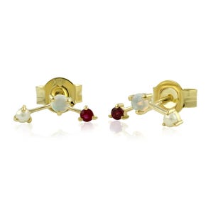 May include: A pair of gold stud earrings, each featuring a cluster design. Each earring has a white pearl, a white opal, and a red ruby gemstone. The earrings are set against a white background.