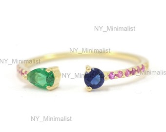 Op de afbeelding: Een gouden ring met een peervormige smaragd edelsteen aan de ene kant en een ronde saffier edelsteen aan de andere kant. De ring is bezet met kleine roze edelstenen.