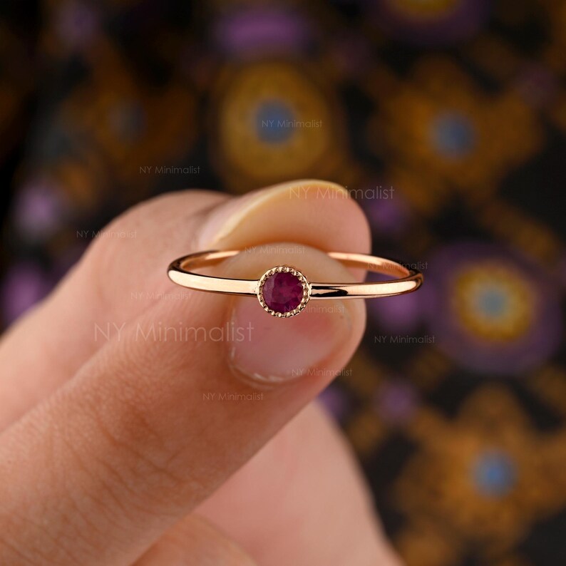 May include: A thin gold ring featuring a single, small, round, deep pink gemstone set in a beaded bezel setting. The band is smooth and polished, showcasing a delicate design that highlights the central stone and its textured metal border.