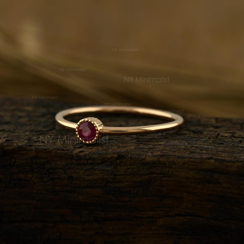 May include: A thin gold ring featuring a small, round, deep red ruby gemstone set in a delicate, textured bezel. The band is smooth and polished, showcasing a simple design that highlights the vibrant color of the central stone.