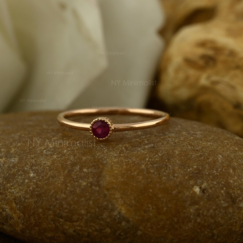 May include: A thin rose gold ring featuring a single, small, round, deep red ruby gemstone set in a bezel setting. The metal band is smooth and circular, showcasing a delicate design with a polished finish that reflects light on the surface.