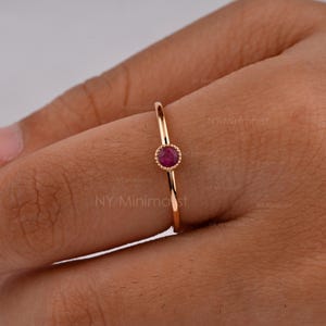 May include: A thin gold ring featuring a single, small, round, deep pink ruby gemstone set in a beaded bezel setting. The band is smooth and polished, showcasing a delicate design that sits centered on the finger with a vibrant gemstone.