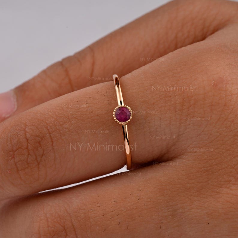 May include: A thin gold ring featuring a single, small, round, deep pink ruby gemstone set in a beaded bezel setting. The band is smooth and polished, showcasing a delicate design that sits centered on the finger with a vibrant gemstone.